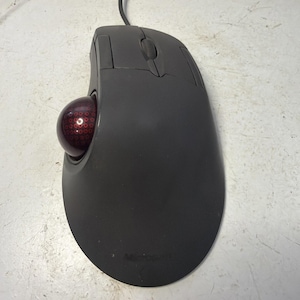 Puede incluir: Un ratón trackball gris oscuro con una bola roja y una rueda de desplazamiento. El ratón tiene una conexión por cable y la marca "Microsoft" es visible en la parte inferior.