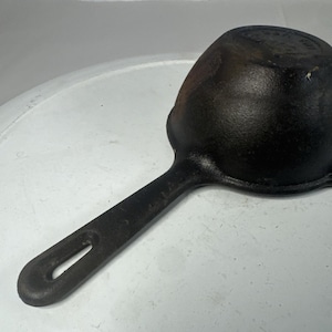 May include: A black cast iron skillet with a round bowl and a long, straight handle featuring an elongated hole at the end. The exterior surface of the pan shows a textured, dark finish with subtle signs of wear and a slightly rusted appearance.
