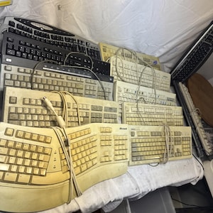 以下が含まれることがあります： ベージュ、黒、グレーの様々なコンピューターキーボードのコレクション。QWERTY配列の有線モデルが複数含まれており、曲線的なMicrosoftの人間工学に基づいたキーボードや、標準的な長方形のDell製ユニットが複数並んでいます。