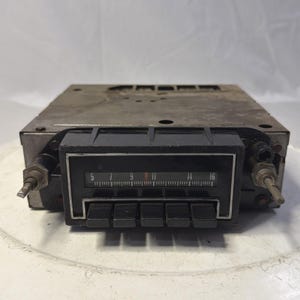 May include: Vintage car radio with a black faceplate and silver metal casing. The radio features a tuning dial with numbers from 5 to 16 and several black push buttons. Knobs and other components are visible on the sides.