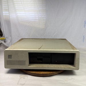 May include: A vintage off-white IBM computer. The front panel has a vertical vent, a floppy disk drive, and a black disk drive. The IBM logo is visible on the left. The computer is on a wooden base.