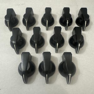 May include: A collection of twelve black control knobs with a silver accent line. The knobs have a rounded base and a rectangular top, likely for use on electronic equipment or musical instruments.