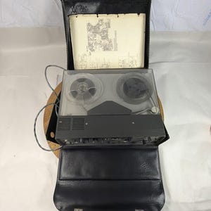 May include: Vintage reel-to-reel tape recorder in a black leather case. The recorder has clear plastic reels and a gray body. A schematic diagram is visible inside the open case. The case is open, revealing the recorder and its components.
