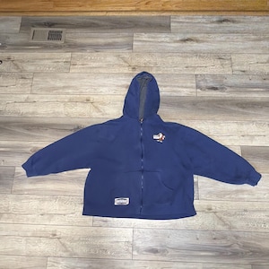 Könnte beinhalten: Ein marineblauer Kapuzenpullover mit grauem Kapuzenfutter und einem kleinen gestickten Design auf der Brust. Der Kapuzenpullover hat eine Kängurutasche und ein kleines Etikett unten. Das Kleidungsstück liegt auf einer Holzoberfläche.