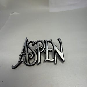 May include: Silver-colored metal sign with the word "ASPEN" in a stylized font. The letters are connected and have a slightly raised, three-dimensional appearance. The sign is likely a decorative item.