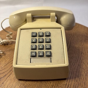 May include: Cream-colored vintage telephone with a rectangular base and a handset. The phone features a keypad with black number and letter keys. The phone is on a wooden surface.