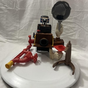 May include: A whimsical, multi-piece sculpture featuring a vintage-style camera with a wooden body, a red fire hydrant, and a flash held by a hand. The sculpture has a brown horse-shaped stand and a red tray.