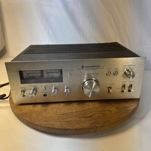 Könnte beinhalten: Ein silberner Kenwood Stereo-Vollverstärker Modell KA-5500 mit einer Frontplatte aus gebürstetem Metall, zwei analogen Leistungsanzeigen, einem großen zentralen Lautstärkeregler, mehreren kleineren Drehreglern, Kippschaltern und einem schwarzen Gehäuse.