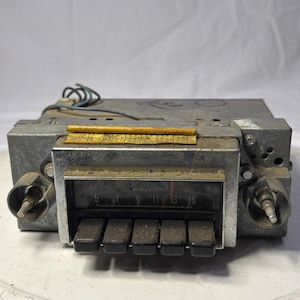 May include: Vintage Philco car radio with a silver faceplate and black buttons. The radio dial displays numbers 5, 7, 9, 11, 13, and 15. The radio has two knobs and a series of wires extending from the top.