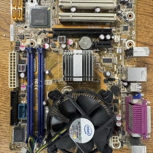 Puede incluir: Una placa base de escritorio Intel de color marrón que cuenta con un ventilador de refrigeración negro con el logotipo de Intel, dos ranuras RAM azules, dos ranuras PCI blancas, un disipador de calor de metal plateado y varios puertos integrados.