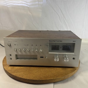 May include: Silver and wood-tone stereo eight-track deck. The face has buttons, dials, and meters. The top is wood-grained. The model number is 3331. The deck is on a round wooden surface.