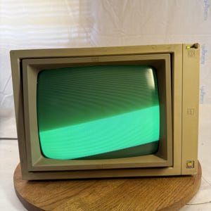 Peut inclure: Un ancien moniteur d'ordinateur beige avec un écran vert. Le moniteur a un écran carré et un cadre épais. Il est posé sur une base en bois.