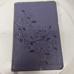 May include: A purple book with a decorative floral design on the cover. The design features swirling vines and leaves in a darker shade of purple. The book has a slightly worn appearance.