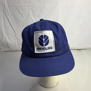 Op de afbeelding: Een blauwe baseballpet met een gebogen klep. De pet heeft een wit vierkant embleem met het New Holland-logo in blauw. De tekst "NEW HOLLAND" staat eronder. Gemaakt van duurzame stof.