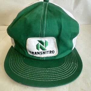 Puede incluir: Gorra de camionero verde y blanca con visera curva. El panel frontal es verde con un parche blanco con la palabra "TRANSNITRO" y un logotipo verde y blanco. La parte trasera de la gorra es de malla blanca.