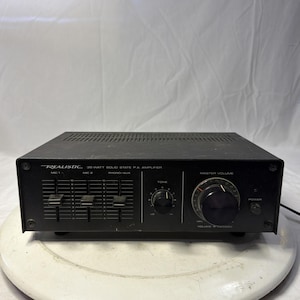Puede incluir: Amplificador PA de estado sólido Realistic negro de 35 vatios. El panel frontal presenta controles deslizantes para MIC 1 y MIC 2, un control de tono y una perilla de volumen maestro. El botón de encendido está a la derecha.