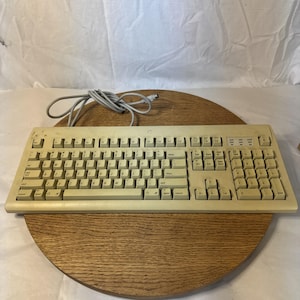 Pode incluir: Um teclado de computador vintage bege com layout QWERTY, teclado numérico e um cabo cinza em espiral que se estende pela parte traseira. O teclado possui teclas de função, teclas de comando e um arranjo alfanumérico padrão retangular.