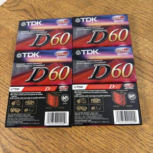 Puede incluir: Seis cintas de casete de audio TDK D60. El embalaje es rojo y negro con el logotipo de TDK y "D60" en letras plateadas grandes. También es visible el texto "Dynamic Performance". Las cintas están dispuestas sobre una superficie de madera.