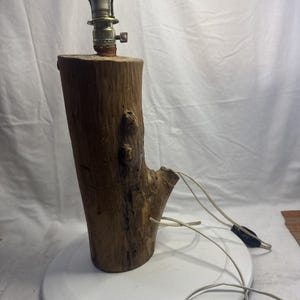 May include: A rustic wooden table lamp. The base is a section of a tree trunk with a natural, textured surface. The lamp features a brass-colored socket and a white electrical cord. The lamp is designed to provide ambient lighting.