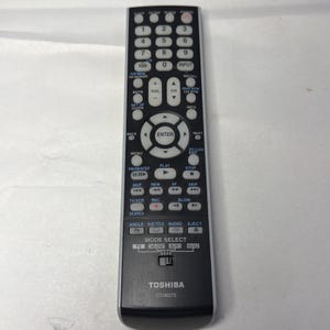 Puede incluir: Un mando a distancia negro Toshiba CT-90275 con botones blancos. El mando a distancia tiene una variedad de botones para encendido, volumen, canal y navegación por el menú. La palabra "TOSHIBA" está impresa en la parte inferior.