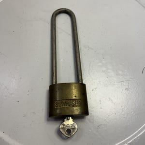 Puede incluir: Un candado de latón con un grillete de plata y una llave. El candado tiene la palabra "SLAYMAKER" impresa en él.