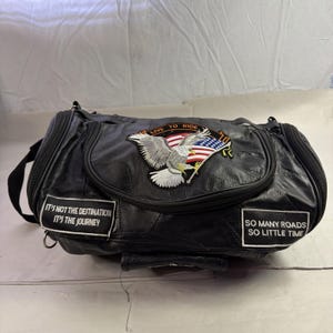 May include: Black leather duffel bag with a patchwork design. Features embroidered patches with an eagle, American flag, and text that reads "LIVE TO RIDE", "IT'S NOT THE DESTINATION IT'S THE JOURNEY", and "SO MANY ROADS SO LITTLE TIME". Includes a black shoulder strap.