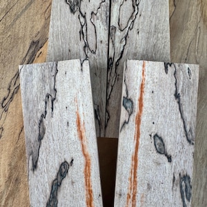 May include: A collection of light-colored wood pieces with unique black and orange veining patterns. The wood has a natural, textured appearance, with the grain visible. The pieces are arranged to showcase the intricate designs.