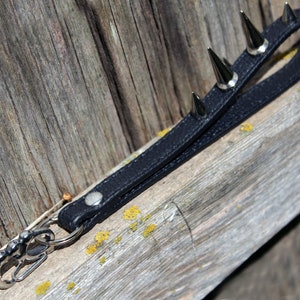 May include: Black leather keychain wrist strap with silver-colored metal spikes and a silver-colored metal clasp. The strap is attached to a wooden surface.