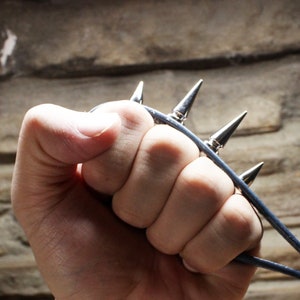 May include: A silver-toned claw weapon with four sharp spikes, held in a clenched fist. The spikes are attached to a dark blue leather strap. The background is a textured, light brown stone wall.