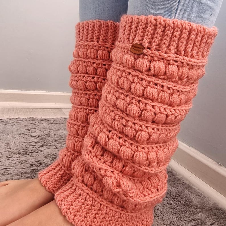 K&ouml;nnte beinhalten: Ein Paar korallenrosa geh&auml;kelte Beinstulpen mit einem strukturierten Noppenmuster. Die Beinstulpen haben ein braunes Etikett mit der Aufschrift "Stephanie Threads".