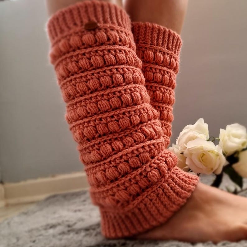 K&ouml;nnte beinhalten: Ein Paar korallenfarbene geh&auml;kelte Beinstulpen mit einem strukturierten Noppenmuster. Die Beinstulpen werden an den Beinen einer Person getragen und reichen bis knapp unter das Knie.