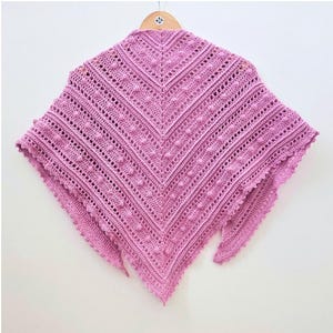 Puede incluir: Un chal de ganchillo hecho a mano, de color lila claro, con un diseño calado texturizado. El chal triangular tiene un borde festoneado y una forma de cuello en V. El hilo tiene un acabado suave y mate. Se muestra en una percha de madera.