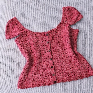 Puede incluir: Un top de ganchillo rojo con mangas cortas y botones en la parte delantera. El top tiene un patrón floral y un dobladillo festoneado. El diseño de hombros descubiertos añade un toque de elegancia. El top está hecho de hilo suave y texturizado.