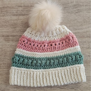 Puede incluir: Un gorro de ganchillo hecho a mano con un pompón color crema. El gorro presenta rayas alternas de crema, rosa y verde azulado. El borde inferior es acanalado y el gorro está hecho de hilo suave.