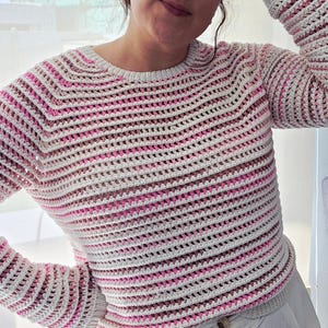 Puede incluir: Un suéter de crochet hecho a mano con un patrón de rayas en blanco, rosa y gris. El suéter tiene un cuello redondo y mangas largas. El diseño de tejido abierto es visible en toda la prenda.