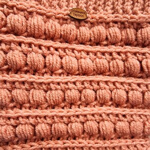 K&ouml;nnte beinhalten: Nahaufnahme eines geh&auml;kelten Stoffes mit einer strukturierten, unebenen Oberfl&auml;che. Der Stoff ist in einem hellen Pfirsich-Ton gehalten. Ein kleines Holzschild mit dem Text "STEPHANIE TEJIDOS" ist am Stoff befestigt.