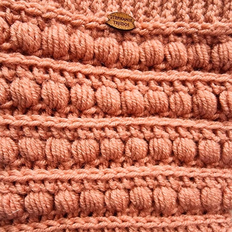 K&ouml;nnte beinhalten: Nahaufnahme eines geh&auml;kelten Stoffes mit einer strukturierten, unebenen Oberfl&auml;che. Der Stoff ist in einem hellen Pfirsich-Ton gehalten. Ein kleines Holzschild mit dem Text "STEPHANIE TEJIDOS" ist am Stoff befestigt.