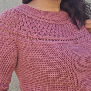 Puede incluir: Un suéter de color rosa empolvado con un patrón de punto texturizado. El cuello presenta un diseño calado decorativo. La prenda de manga larga está hecha de un material suave, posiblemente lana o algodón.