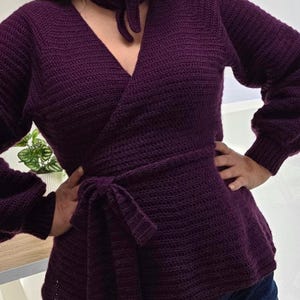 Puede incluir: Un suéter cruzado de manga larga en color morado oscuro con un patrón de tejido de ganchillo texturizado, escote en V y un cierre de lazo a juego en la cintura. La prenda incluye un pañuelo de cuello coordinado anudado al frente y puños acanalados.