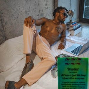 Puede incluir: Una pastilla de jabón verde y negro con el texto "Drakkar Goat Milk Soap" y "All Natural Goat Milk Soap infused with a sea sponge". El jabón está sobre una superficie blanca con una persona en el fondo.