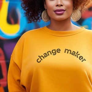 May include: A woman with curly brown hair is wearing a yellow sweatshirt with the text "change maker" printed on the front. She is standing in front of a colourful graffiti wall.
