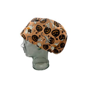 May include: A tan Halloween-themed cap featuring a repeating pattern of black jack-o'-lanterns and cartoon characters. The cap is made of fabric and has a rounded shape. The characters include a bear, a tiger, and a pig.