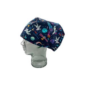 May include: A navy blue surgical scrub cap featuring a colorful space-themed pattern. The design includes astronauts, planets, rockets, and other celestial elements. The cap is made of a soft, flexible material.