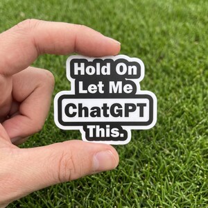 May include: A die-cut sticker with a white background and a thick black border. The sticker features the black text "Hold On Let Me ChatGPT This." arranged in three lines within a rectangular shape with rounded corners and a central protrusion.