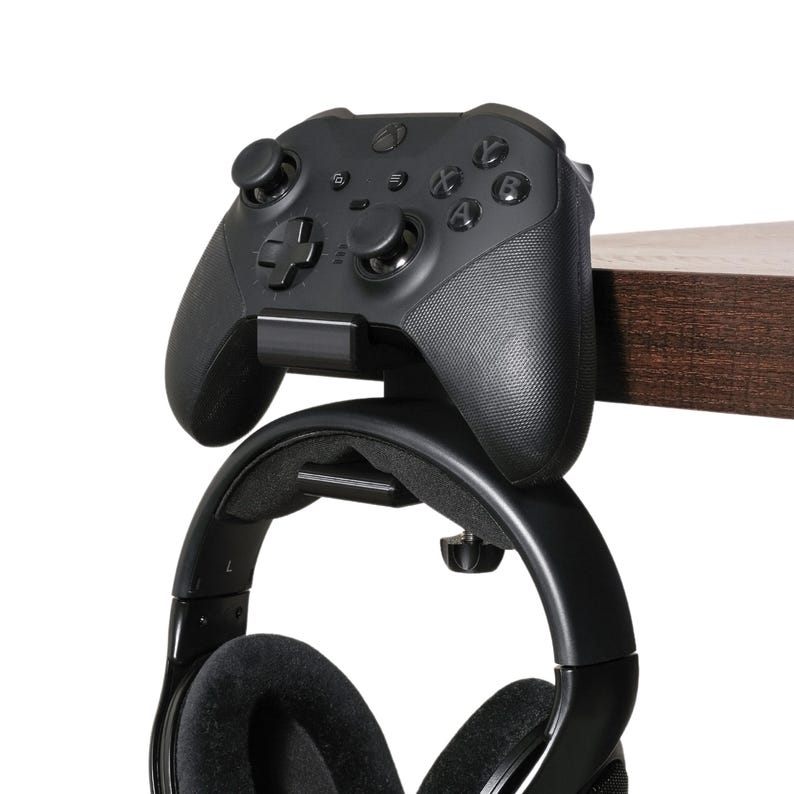 Xbox One controller sits upright on the controller and headphone holder clamped to the side of a desk. A pair of headphones hangs below the controller on a hook attached to the desk-mount holder.