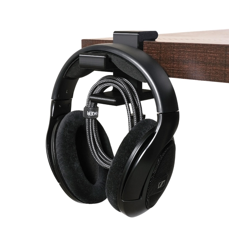 A pair of headphones hangs from the desk-mount holder clamped to the side of a desk. A charging cable is wrapped neatly and also hangs from the desk-mount holder.