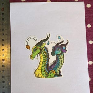 Peut inclure: Un autocollant d&eacute;coup&eacute; repr&eacute;sentant deux t&ecirc;tes de dragon stylis&eacute;es c&ocirc;te &agrave; c&ocirc;te. Le dragon de gauche est vert et jaune avec une antenne termin&eacute;e par une feuille, tandis que celui de droite est bleu et violet. Une r&egrave;gle indique 9 cm de hauteur.