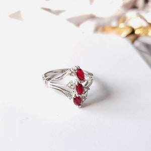 May include: A silver ring with three red oval-shaped gemstones and smaller clear gemstones. The ring has a delicate, intricate design.