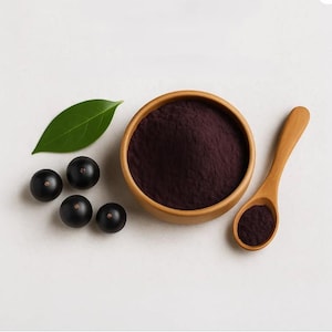 May include: A wooden bowl filled with fine dark purple acai powder, accompanied by a small wooden spoon containing a portion of the powder, four round black acai berries, and a single smooth green leaf arranged on a plain light surface.