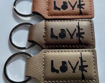 May include: Three leather keychains in shades of brown, each featuring the word "LOVE" with weapon silhouettes. The designs include a pistol, grenade, knife, and rifle. Each keychain has a silver metal ring.